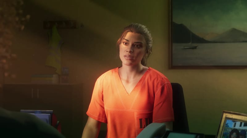 Lucia Caminos, wearing an orange prison shirt, sits at a desk in a dim, warmly lit office with a framed lake painting on the wall behind her.
