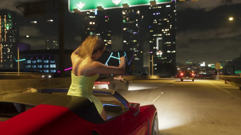 A woman in a pale green dress stands up through the open roof of a red sports car on a night-time motorway, with tall lit-up towers ahead.