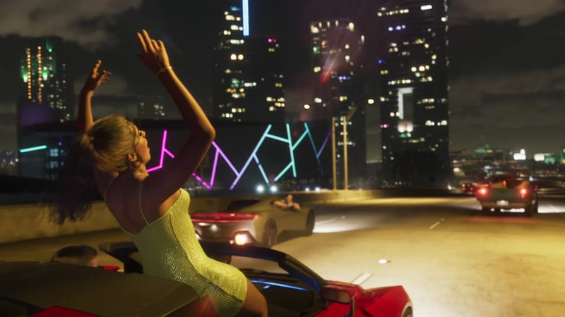 A woman in a sparkly yellow-green dress sits on the back of a red convertible with her arms raised, on a night-time motorway with a lit city skyline behind.