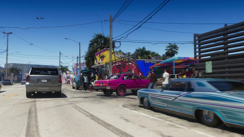 A bright pink lowrider and a blue-striped classic car sit on a sunny Vice City street beside a colourful mural, with a crowd gathered and a helicopter above.