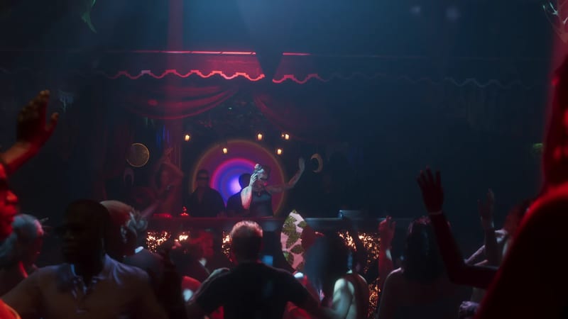 A DJ with one hand raised plays to a packed, dimly lit nightclub crowd, with red and blue lighting and a glowing circular light behind the booth.