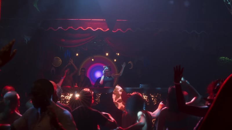 A woman DJ with her hair in buns holds a headphone to her ear behind a booth, with a dancing crowd in the foreground of a dark, red-lit club.
