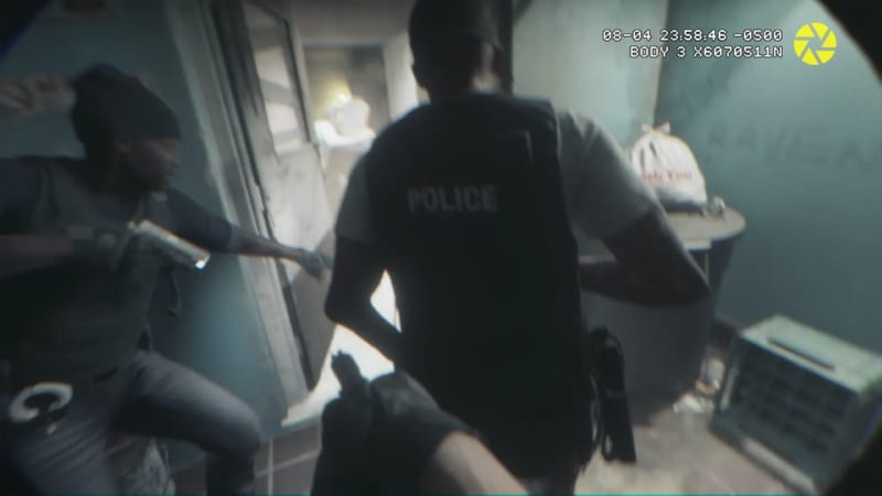 A police officer in a dark vest marked POLICE moves through a dim room as a man in a beanie points a handgun at him, in body camera footage.