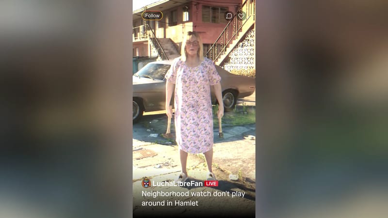 A woman in a pink floral nightdress and glasses stands holding two hammers in front of a battered car and apartment block in Hamlet.