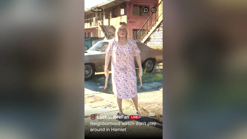 An older blonde woman in glasses and a pink floral dress holds two hammers in a sunlit street outside a run-down red apartment block.