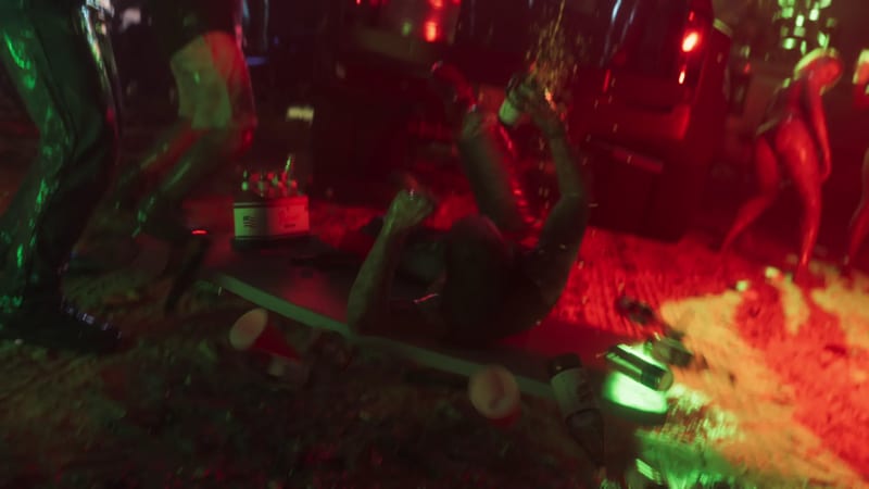 A person lies on their back on the ground at a night-time party, holding up a bottle as drink sprays, with dancers and a dark vehicle nearby.