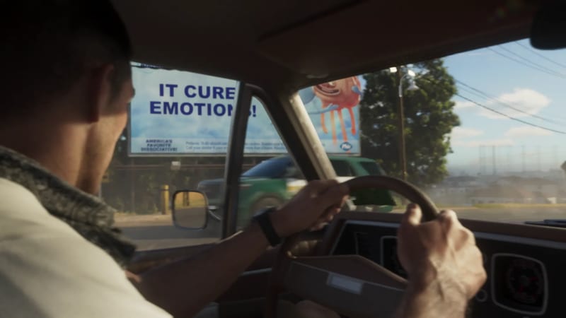 A man in a camouflage-collared jacket drives an old car past a roadside billboard reading "IT CURE EMOTIONS!" on a sunny day.