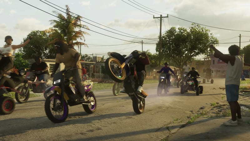 A rider pops a wheelie on a dirt bike on a sunlit residential street as other bikers and quad bikes follow and onlookers cheer.