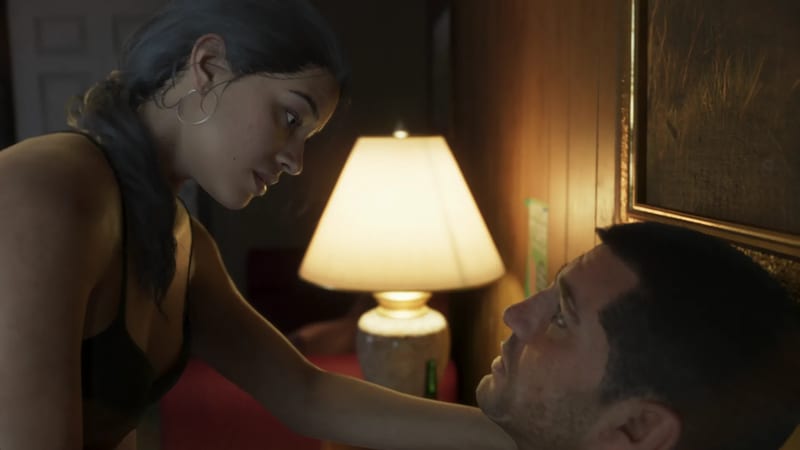 Lucia Caminos leans over Jason Duval, who lies on a bed beside a lit lamp in a warmly lit, wood-panelled room.