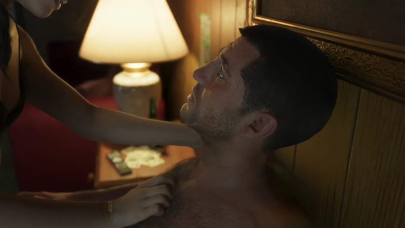 Jason Duval lies shirtless against a wood-panelled headboard as a woman leans over him, in a dimly lit motel room lit by a bedside lamp.