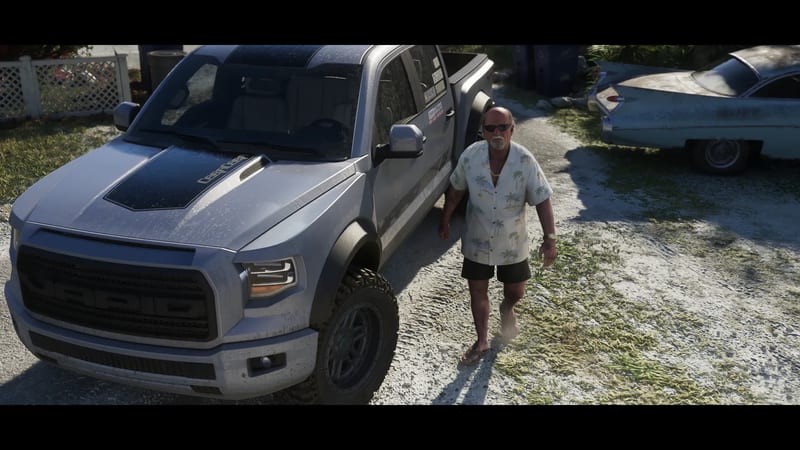 Brian Heder, a bald man in sunglasses and a palm-print shirt, walks barefoot beside a silver Vapid Caracara 4x4 on a sandy yard in bright sun.