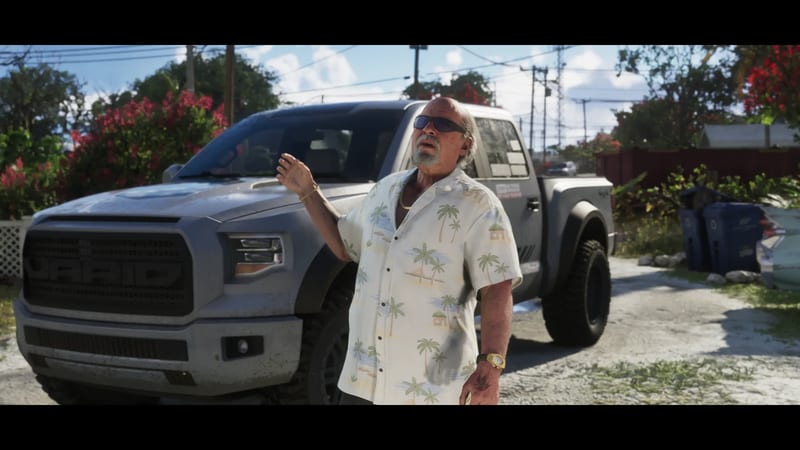 Brian Heder, an older man in sunglasses and a palm-print shirt, raises a hand beside a grey Vapid Caracara 4x4 on a sunny residential driveway.