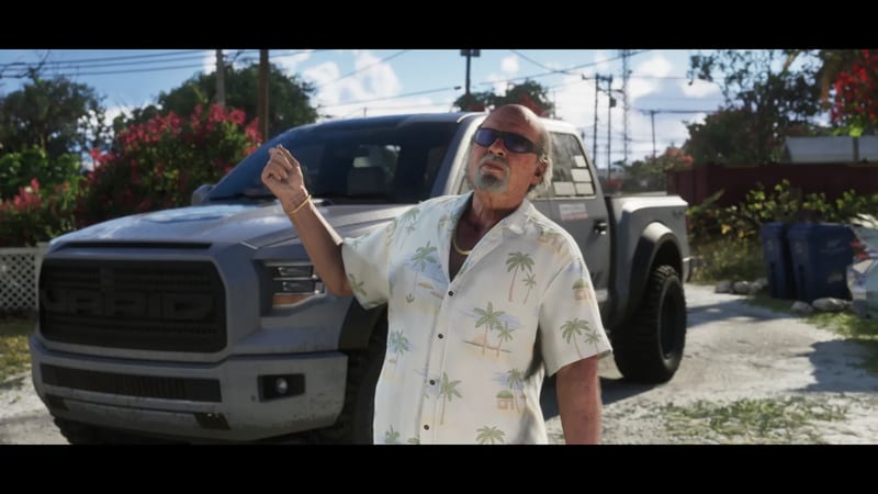 Brian Heder, an older man in sunglasses and a palm-print shirt, gestures in front of a grey Vapid pickup truck on a sunny residential street.
