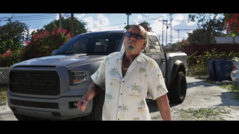 Brian Heder, an older bald man in sunglasses and a palm-print shirt, stands in front of a grey Vapid pickup truck on a sunny residential street.