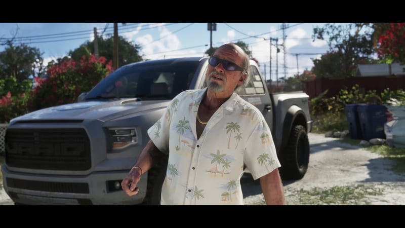 Brian Heder, an older man in sunglasses and a palm-print shirt, stands in front of a grey Vapid pickup truck on a sunny residential street.