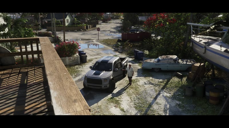 A man in a white shirt stands beside a silver pickup truck on a sandy residential street, seen from a wooden balcony, with a rusty old car and a boat nearby.