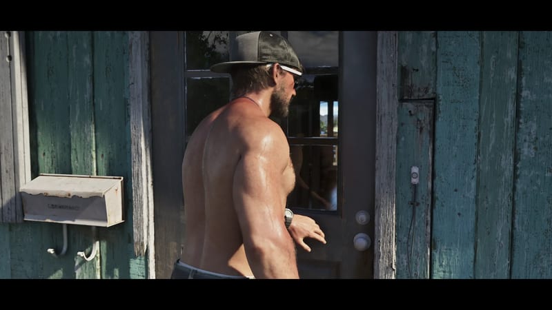 Jason Duval, shirtless in a cap and sunglasses, stands at a glass-panelled door on a weathered teal wooden building in bright sunlight.