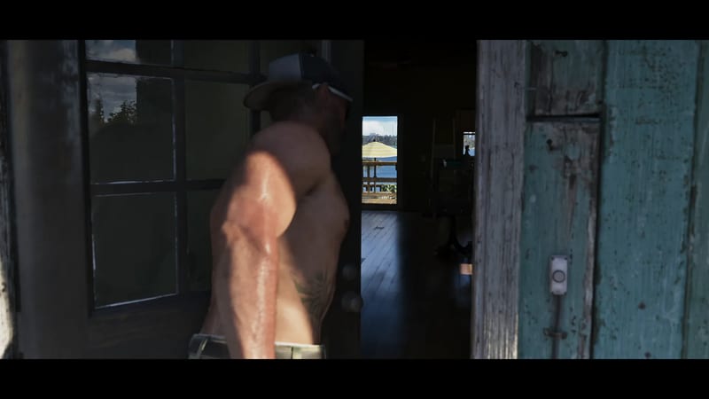 A shirtless man in a cap stands in a doorway of a weathered wooden house, with a deck, water and a yellow striped umbrella visible beyond.