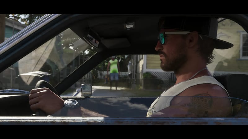 Jason Duval, bearded and in a black cap and sunglasses, drives a car down a sunny residential street, seen in profile through the open window.