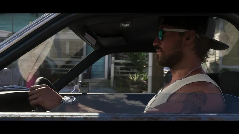 Jason Duval, bearded and in a cap, sunglasses and white vest, drives an old car past pastel wooden houses in daylight.