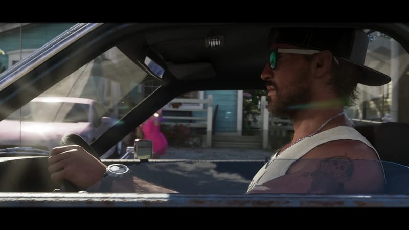 Jason Duval, bearded and wearing a cap and sunglasses, drives an old car past a pastel-blue house on a sunny residential street.