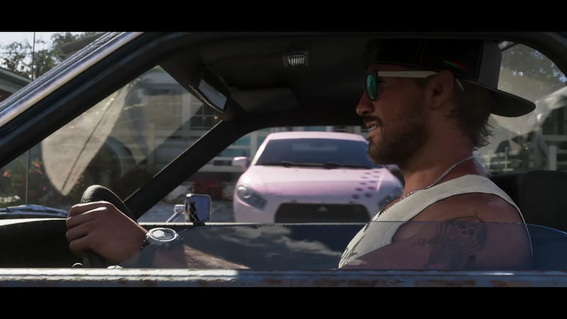 Jason Duval drives a dark car, smiling, in a cap and sunglasses, with a pink hatchback parked behind him on a sunny residential street.