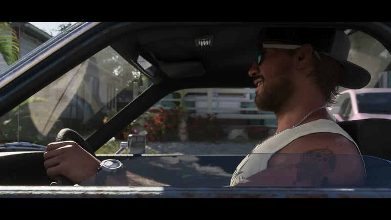 Jason Duval smiles as he drives, one hand on the wheel, past a house with red flowers on a sunny street.