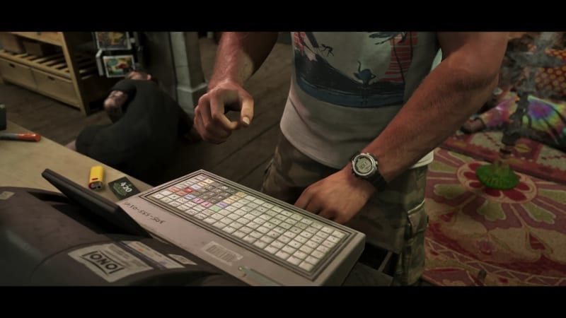 A man in a camouflage shorts and graphic T-shirt types on a cash register keyboard in a cluttered shop while another person lies on the floor behind him.