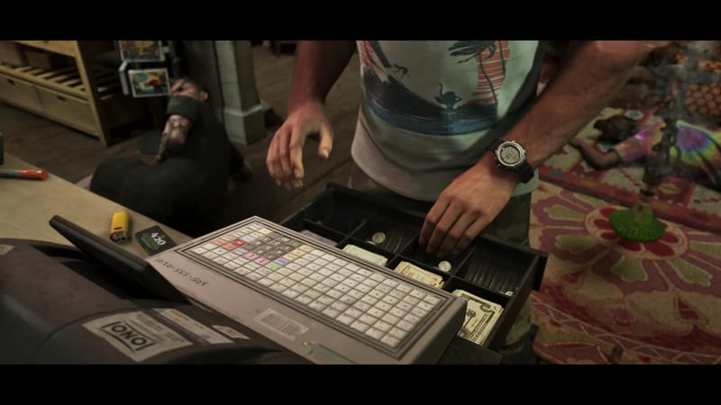 A man in a palm-print T-shirt takes cash from an open till drawer in a shop, while a man sits slumped on the floor behind the counter.