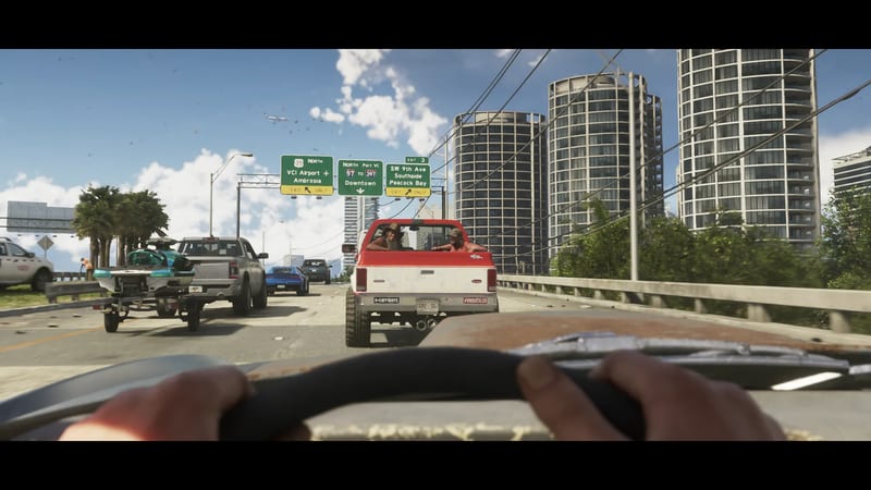 A driver's-eye view of a busy Vice City motorway, with hands on the wheel behind a red and white pickup carrying two men, under overhead green signs.