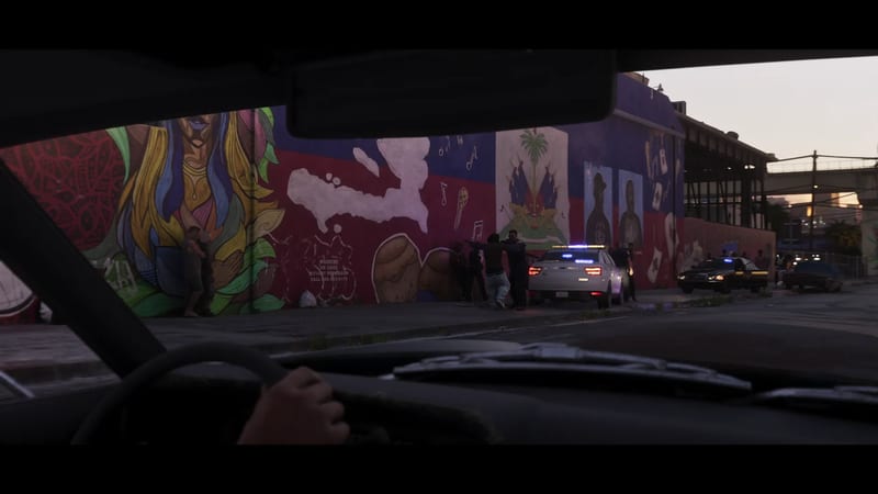 From inside a car, police stop a man against a large mural wall on a street in La Perle at dusk, with a police car's lights flashing.