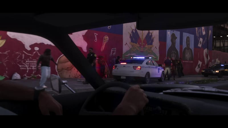 From inside a car at dusk, a driver's hands on the wheel face a police car with flashing lights and officers beside a large mural in La Perle.