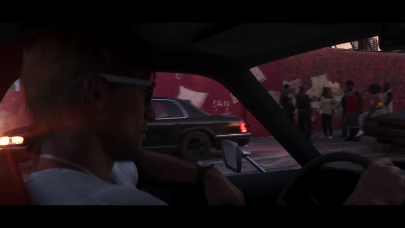 Jason Duval, in sunglasses and a white top, drives at dusk past a black saloon car and a group of people by a red graffiti wall.