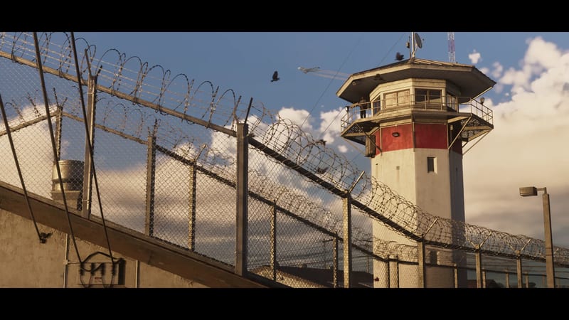 A red and white guard tower stands behind chain-link fences topped with razor wire, with a person on its balcony and a jet in the sky.
