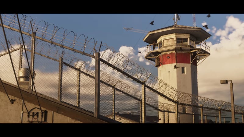 A red and white guard tower stands behind chain-link fences topped with coiled razor wire, with birds flying against a blue sky with clouds.