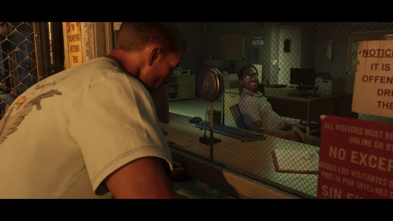 A man in a pale t-shirt leans towards a security window in Leonida Penitentiary, where a smiling man in glasses sits behind a wire-mesh screen.