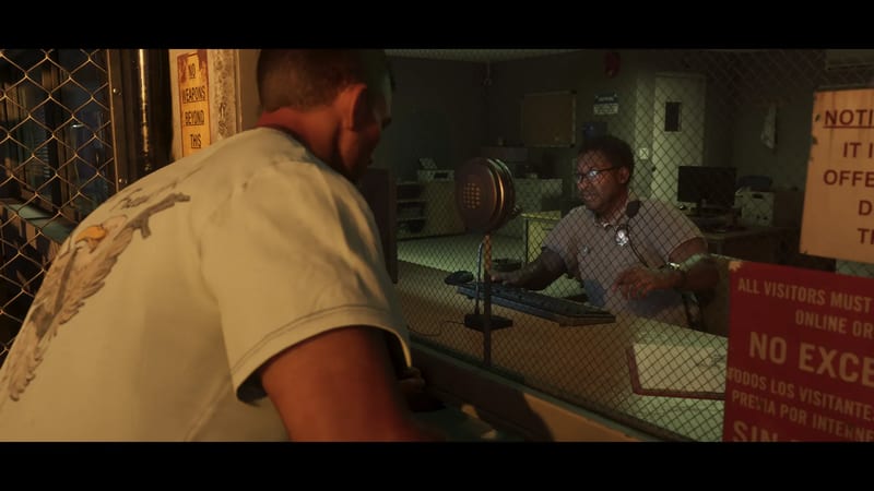 A man in a pale eagle-print T-shirt, seen from behind, speaks to a bespectacled guard through a mesh-covered window in a dimly lit prison office.