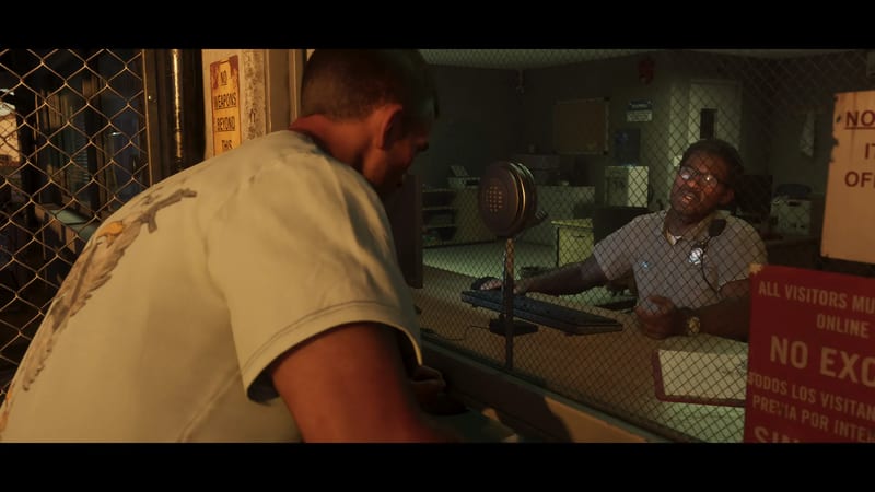 A man in a pale t-shirt leans towards a guard behind a mesh-covered window at a visitor reception desk, lit in warm, dim light.
