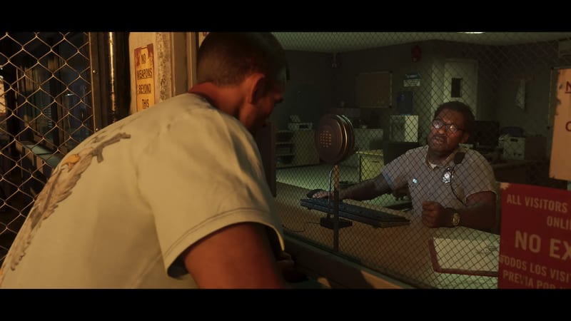 A man in a pale T-shirt leans towards a window to speak to a bespectacled uniformed guard behind a wire mesh screen in a dimly lit reception room.