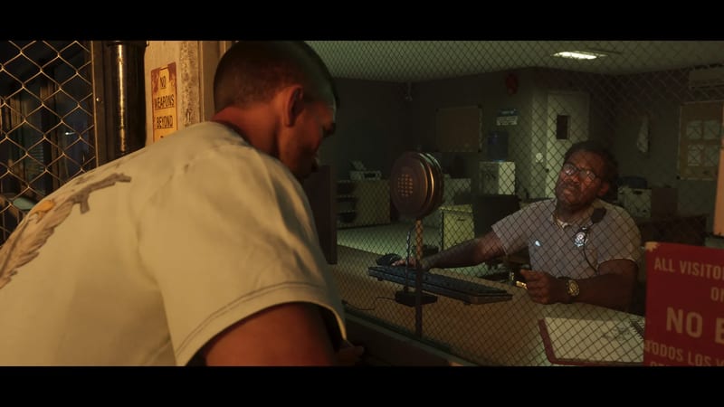 A man in a white t-shirt leans towards a mesh window to speak to a uniformed guard with glasses in a dim, greenish office.