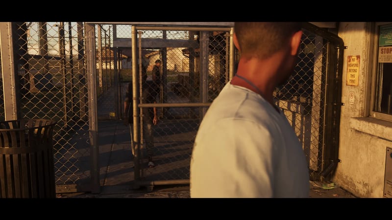 A man in a pale t-shirt, seen from behind, looks through chain-link gates at another man in a fenced walkway in warm evening light.