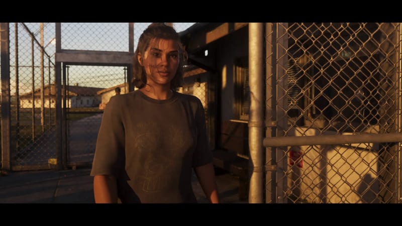 Lucia Caminos stands in a grey T-shirt beside chain-link fences in warm low sunlight at Leonida Penitentiary, with low buildings behind her.