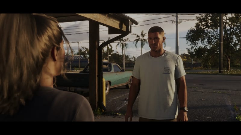 A man in a white Leonida Marine Center T-shirt faces a woman seen from behind, in a car park beside a faded teal pickup.