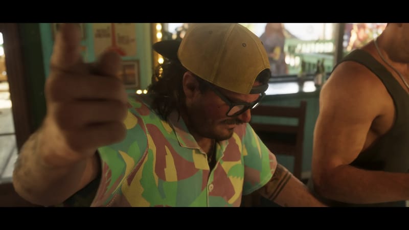 A man in a backwards yellow cap, glasses and a colourful patterned shirt looks down in a bar while another man in a dark vest stands beside him.