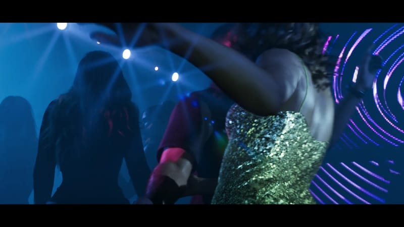 A woman in a green sequinned dress dances with her arm raised, seen from behind, in a blue-lit nightclub crowd with bright spotlights.