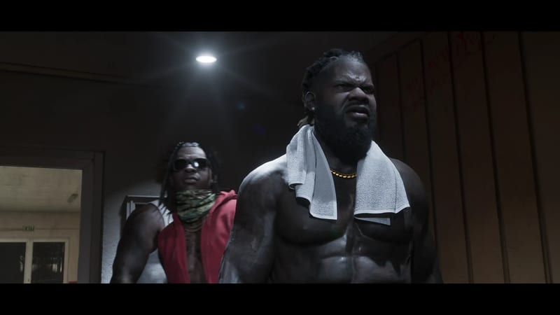 Two muscular, shirtless men stand in a dim room; the front man wears a white towel round his neck and scowls, while the other wears sunglasses and a red vest.