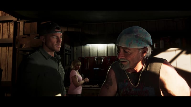 A man in a backwards cap listens to an older bearded man in a blue bandana, with a blonde woman behind them in a dim workshop.