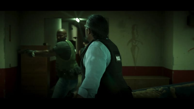 Two police officers in vests move through a dim, green-lit room, one pointing a handgun while the other looks back over his shoulder.