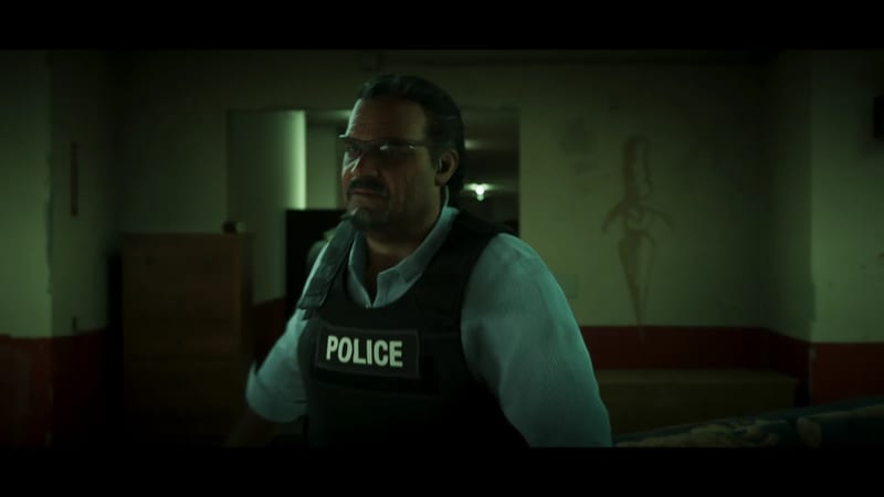 Méndez, a heavyset man with a moustache and glasses, wears a black police vest over a pale blue shirt in a dim, green-lit room.