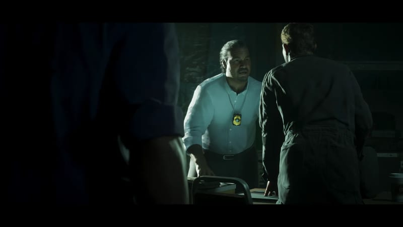 A man in a white shirt with a badge on a chain leans over a desk and speaks to a person in dark overalls, in a dim room.